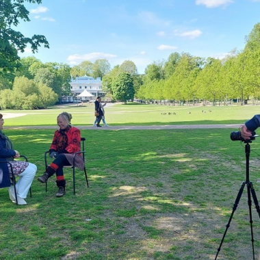 Videokuvaaja taltioi kahden tuoleilla istuvan naisen keskustelua kesäisessä puistossa.